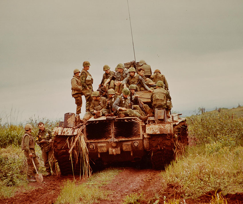 US Army soldiers ride on the back of a tank through a field in South Vietnam