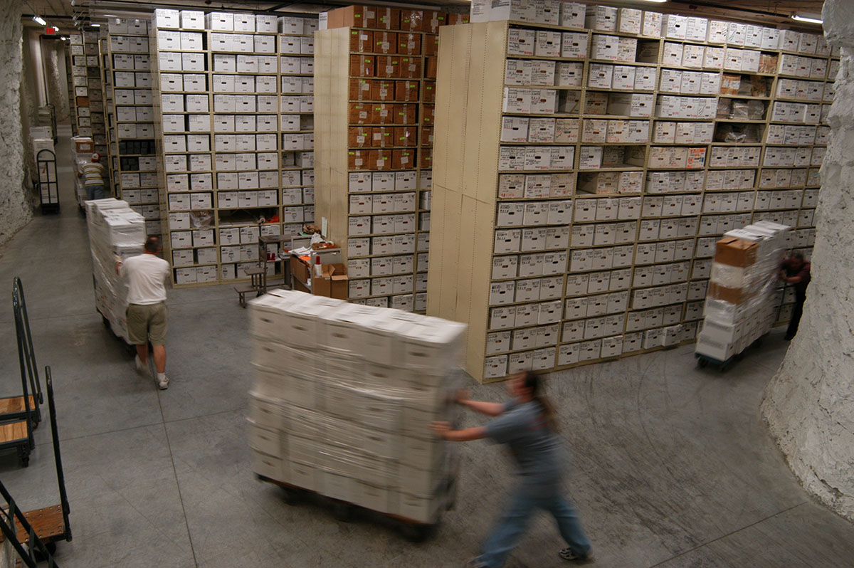 color photograph of stacks with rows of high shelves and staff pushing shrink wrapped boxes of records on pallets