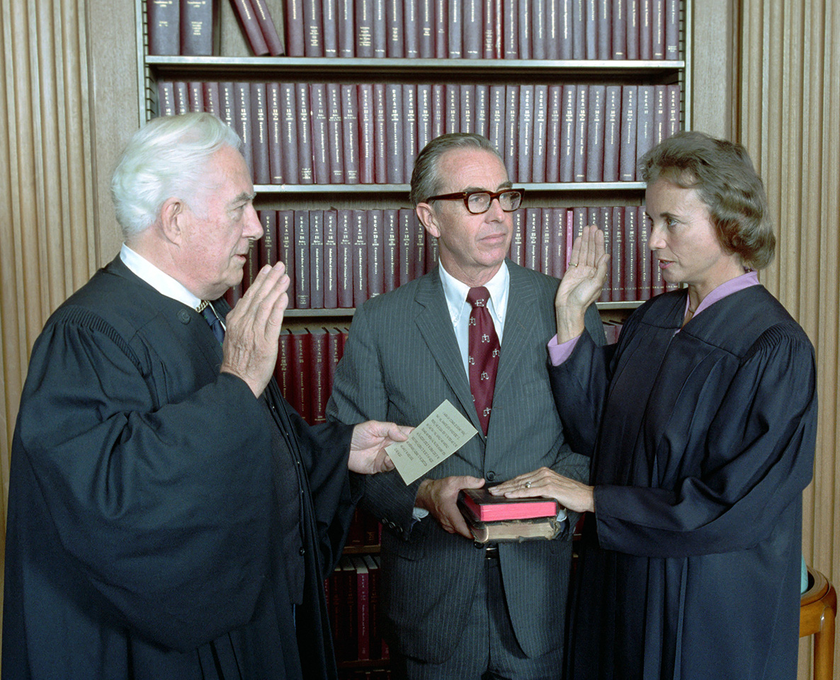 color photograph of a white female judge being sworn in by a white male judge while a white man in a suit holds the books for the swearing in